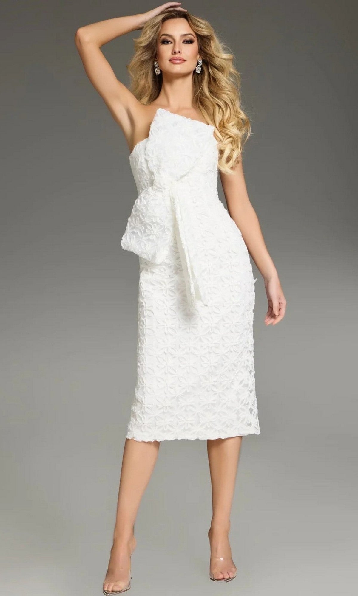 A woman is posing with one arm raised against a plain gray background, wearing the Jovani 39076 Midi Wedding Guest Dress in white—a sleeveless, knee-length design featuring a textured fabric and prominent bow detail.
